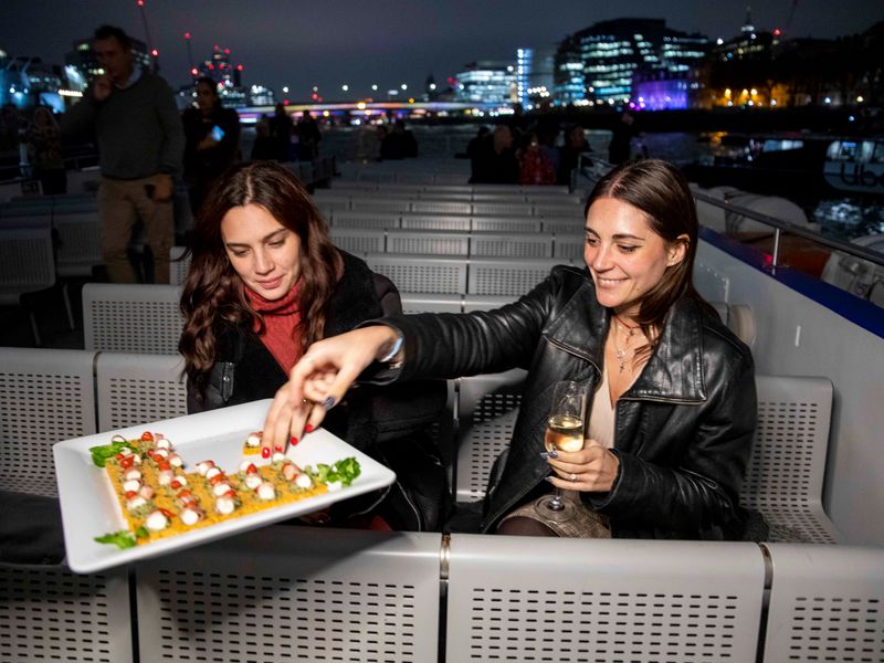 Gäste genießen Snacks und Sekt auf dem Außendeck während einer abendlichen Themsefahrt durch London