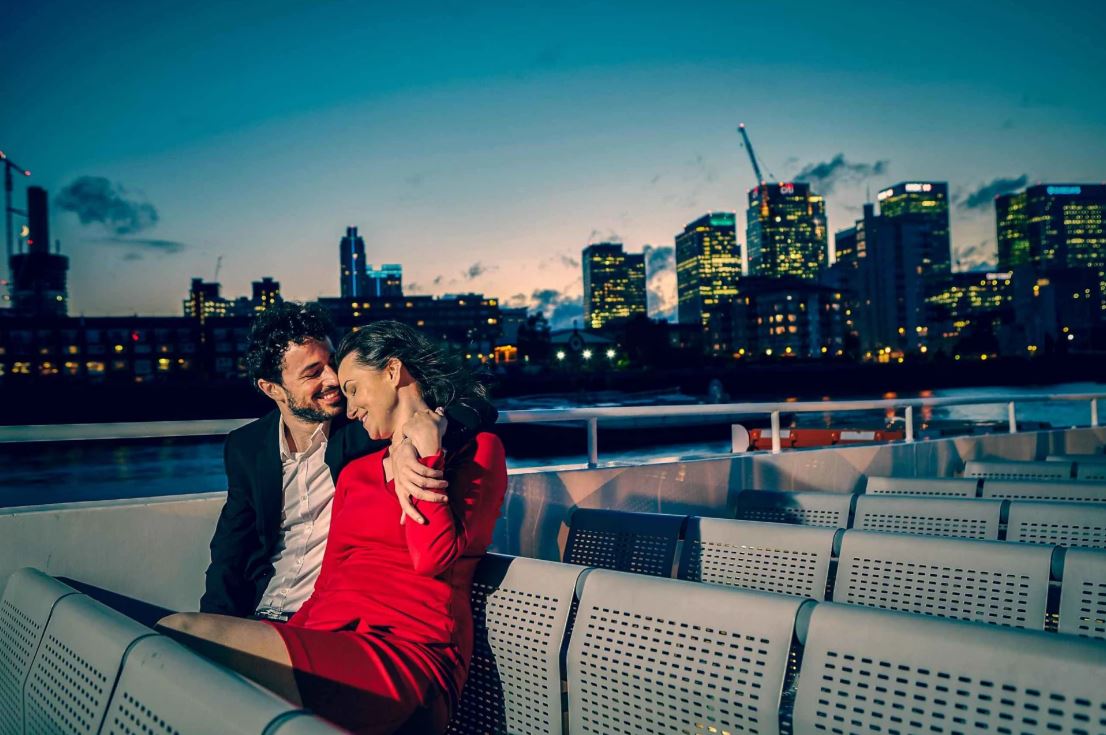 Verliebtes Paar auf Oberdeck der Fähre liegt sich im Arm mit Skyline im Hintergrund bei Nacht in London