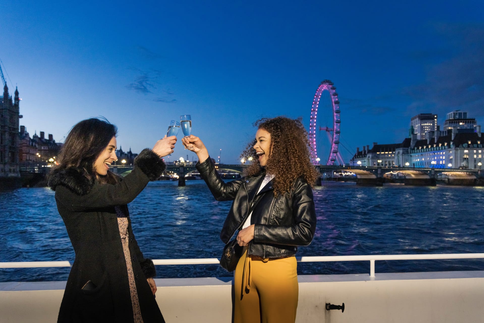 Zwei glückliche Frauen die mit Getränken anstoßen auf Fähre mit beleuchteten London Eye und Skyline im Hintergrund