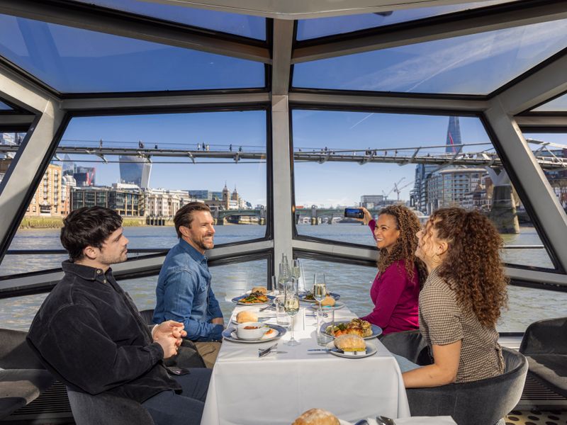 Vier Gäste genießen ein Mittagessen an Bord einer Themsefahrt mit Blick auf die Londoner Skyline