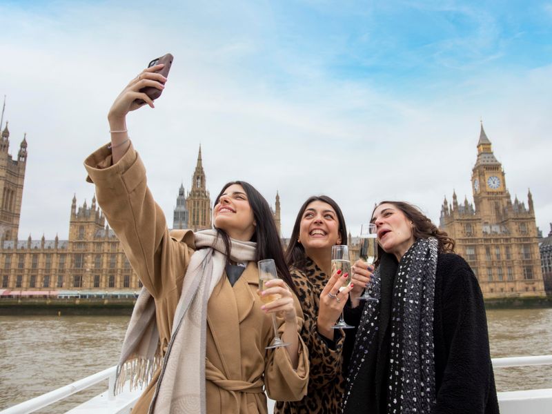 Drei Gäste machen ein Selfie mit Sekt an Bord einer Themsefahrt vor Big Ben und den Houses of Parliament