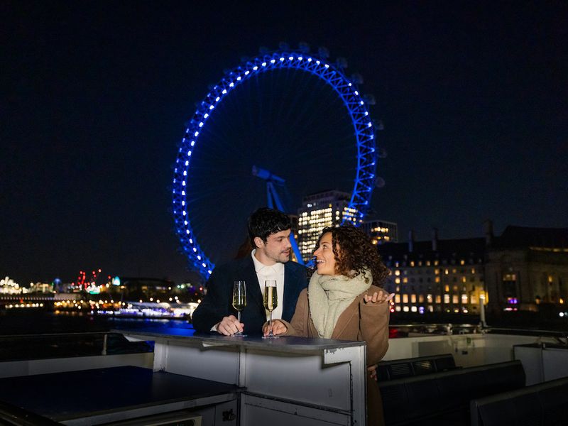 Paar mit Sektgläsern an Deck eines Schiffes vor dem beleuchteten London Eye bei Nacht